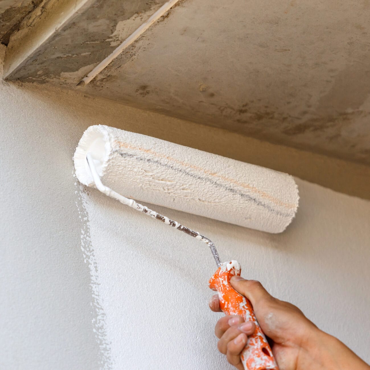 A hand uses a paint roller to apply white paint to a wall near a ceiling.