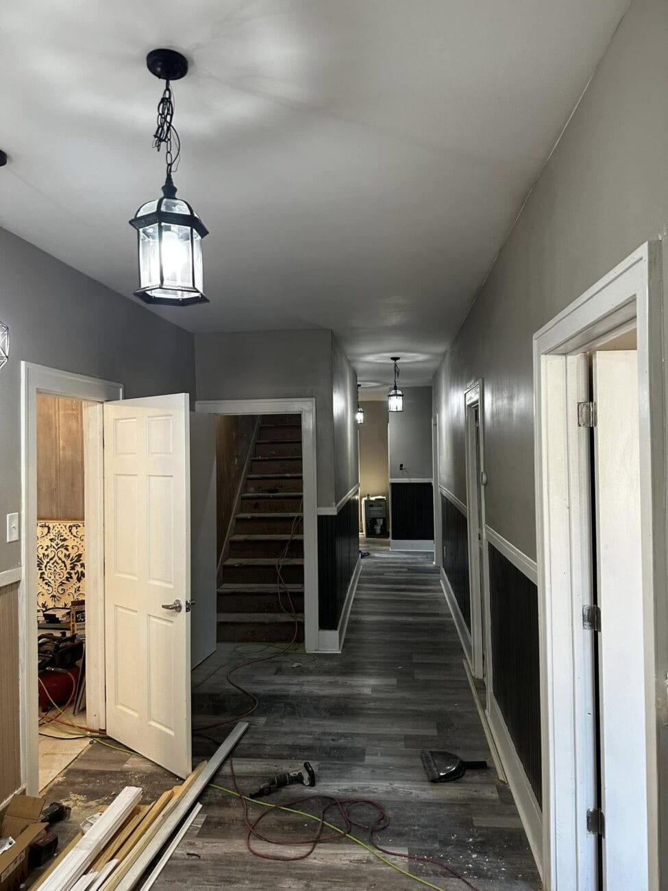 A hallway under renovation with gray walls and flooring, open doors, stairs, and pendant lights. Tools and construction materials are scattered on the floor.